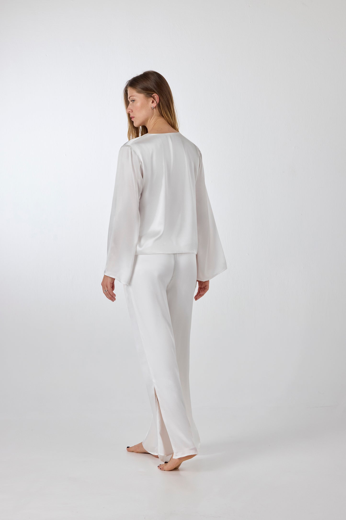 Back view of model wearing A.ROSE Amelia Set in ivory washable silk – adjustable tie‑front long sleeve top paired with wide‑leg pants featuring high side slits and side pockets. Subtly sexy and timeless bridal loungewear/sleepwear, perfect for getting ready or honeymoon mornings.