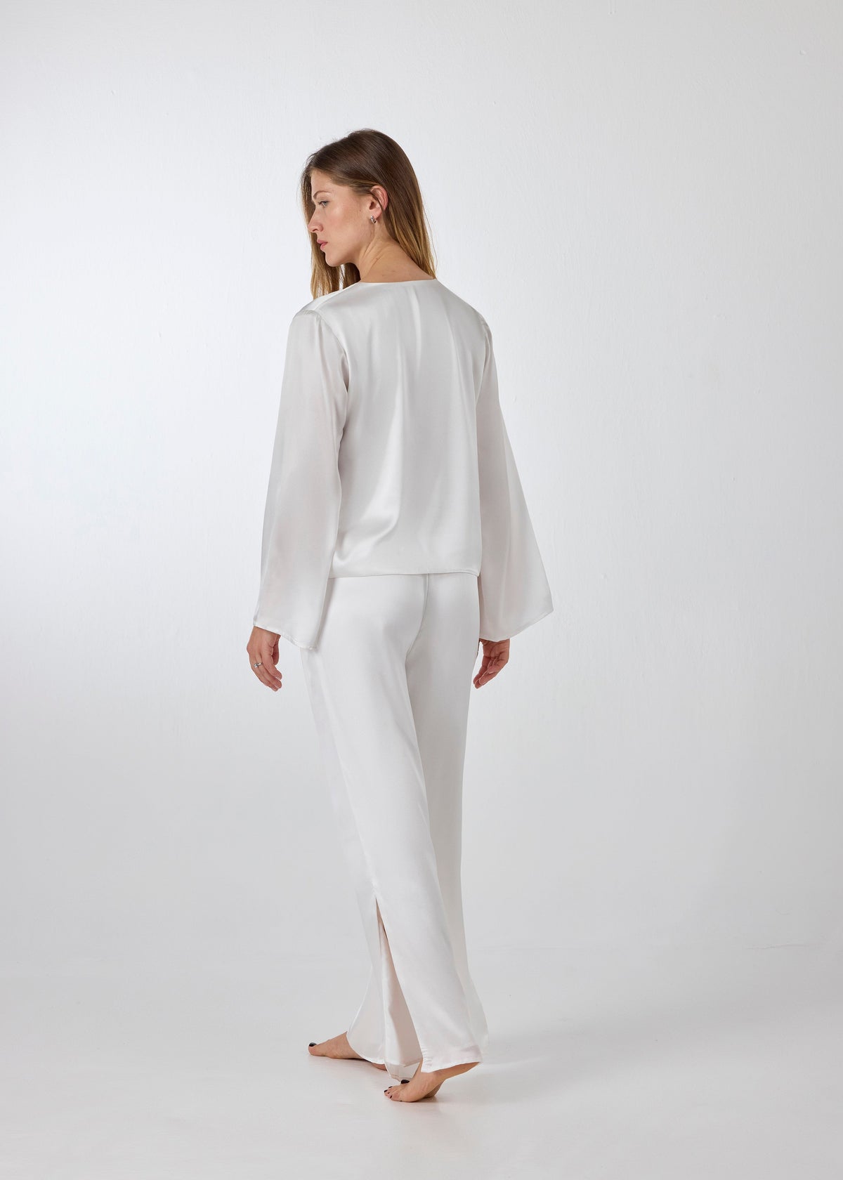 Back view of model wearing A.ROSE Amelia Set in ivory washable silk – adjustable tie‑front long sleeve top paired with wide‑leg pants featuring high side slits and side pockets. Subtly sexy and timeless bridal loungewear/sleepwear, perfect for getting ready or honeymoon mornings.